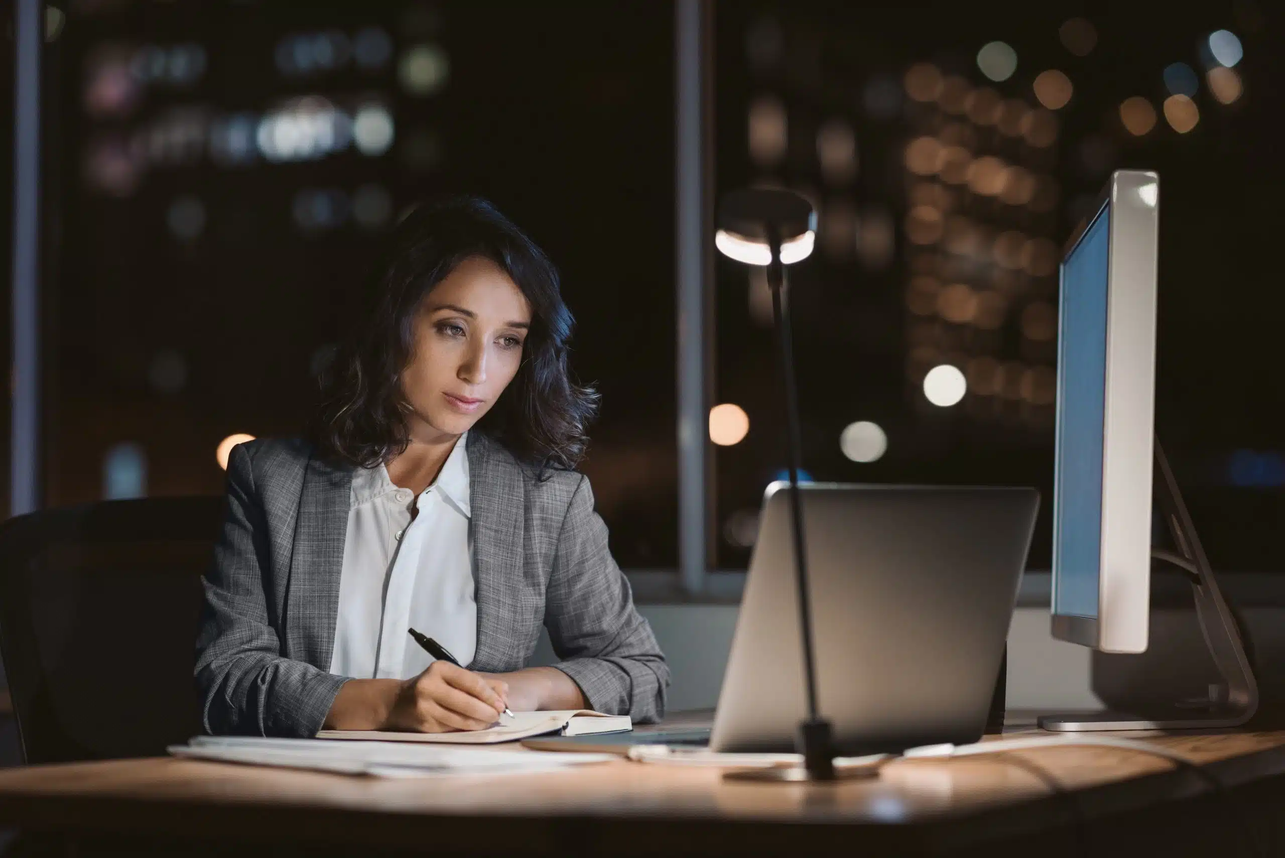 Woman working late at computer, writing notes.