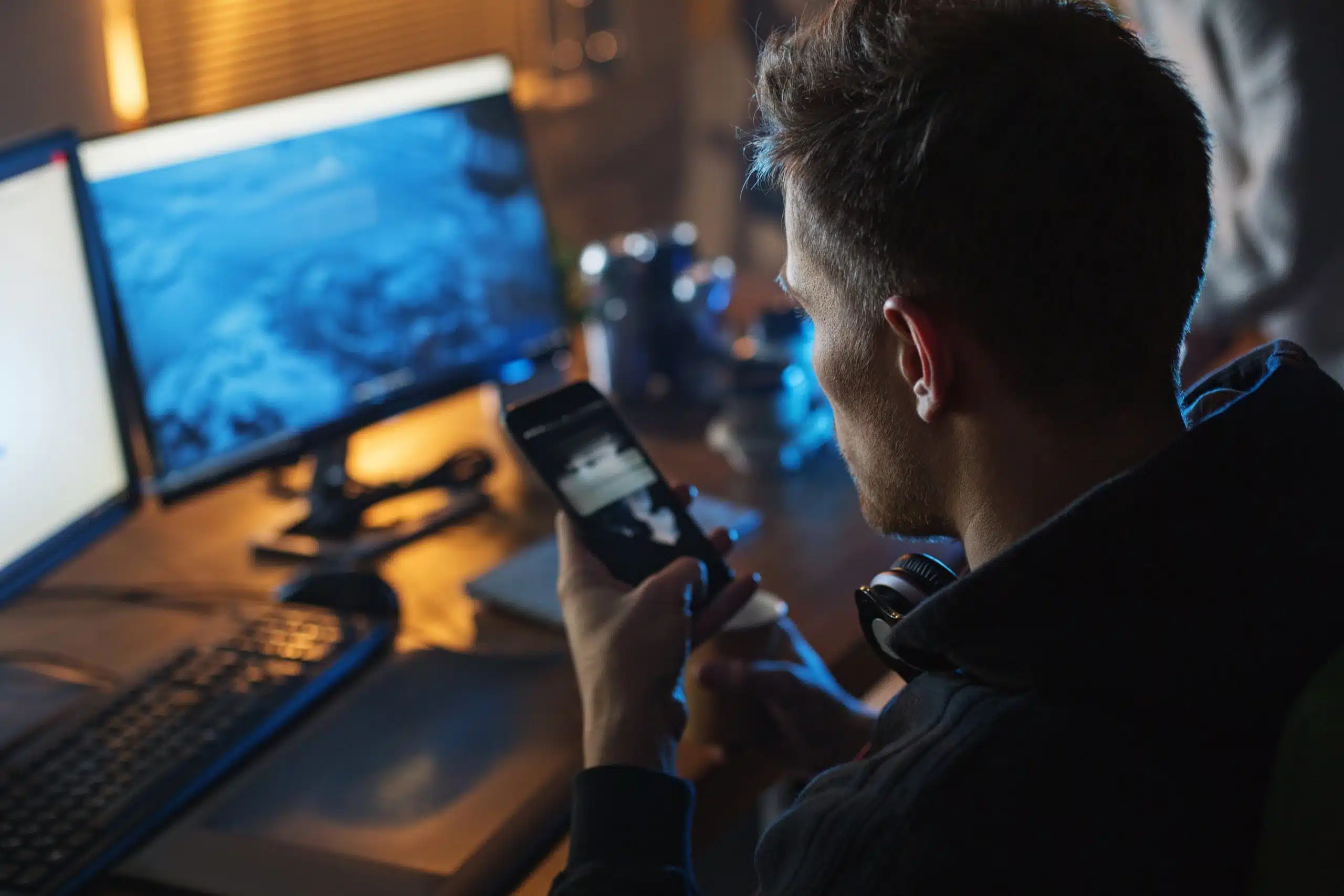 Man using smartphone at desk with computer screens.