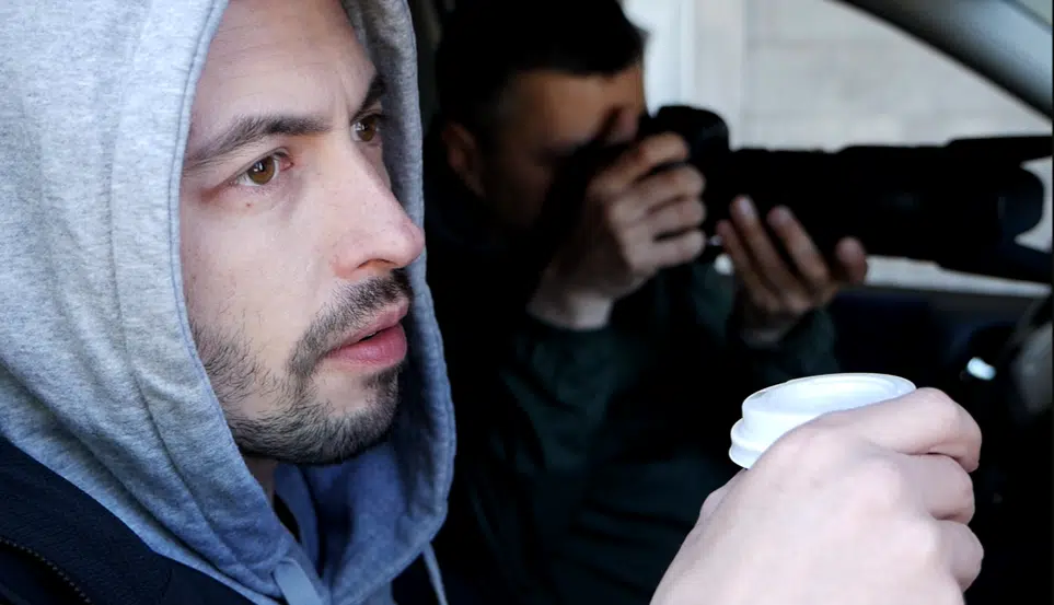 Hooded man holding coffee, photographer in background.