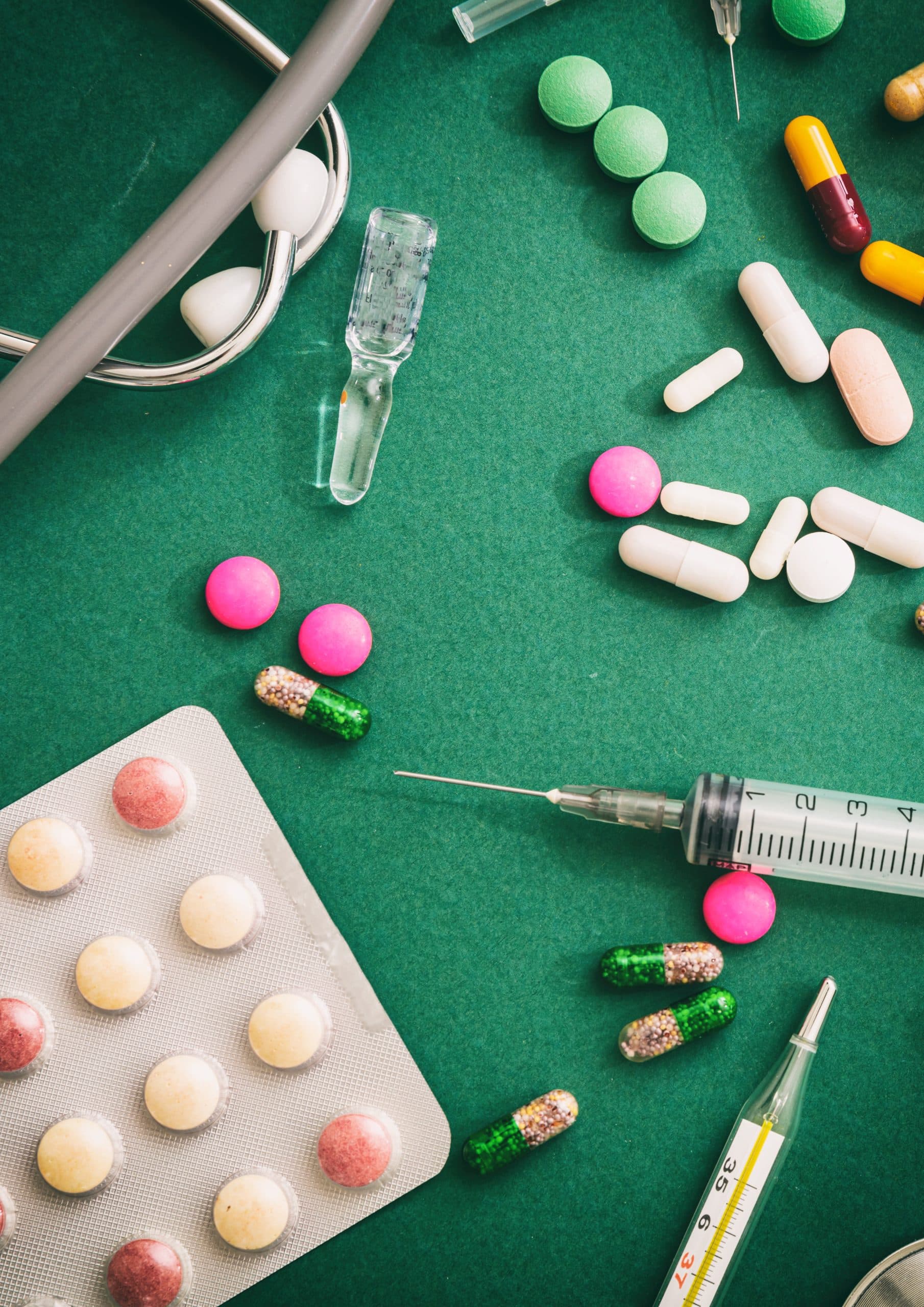 Assorted pills, syringe, and stethoscope on green surface.