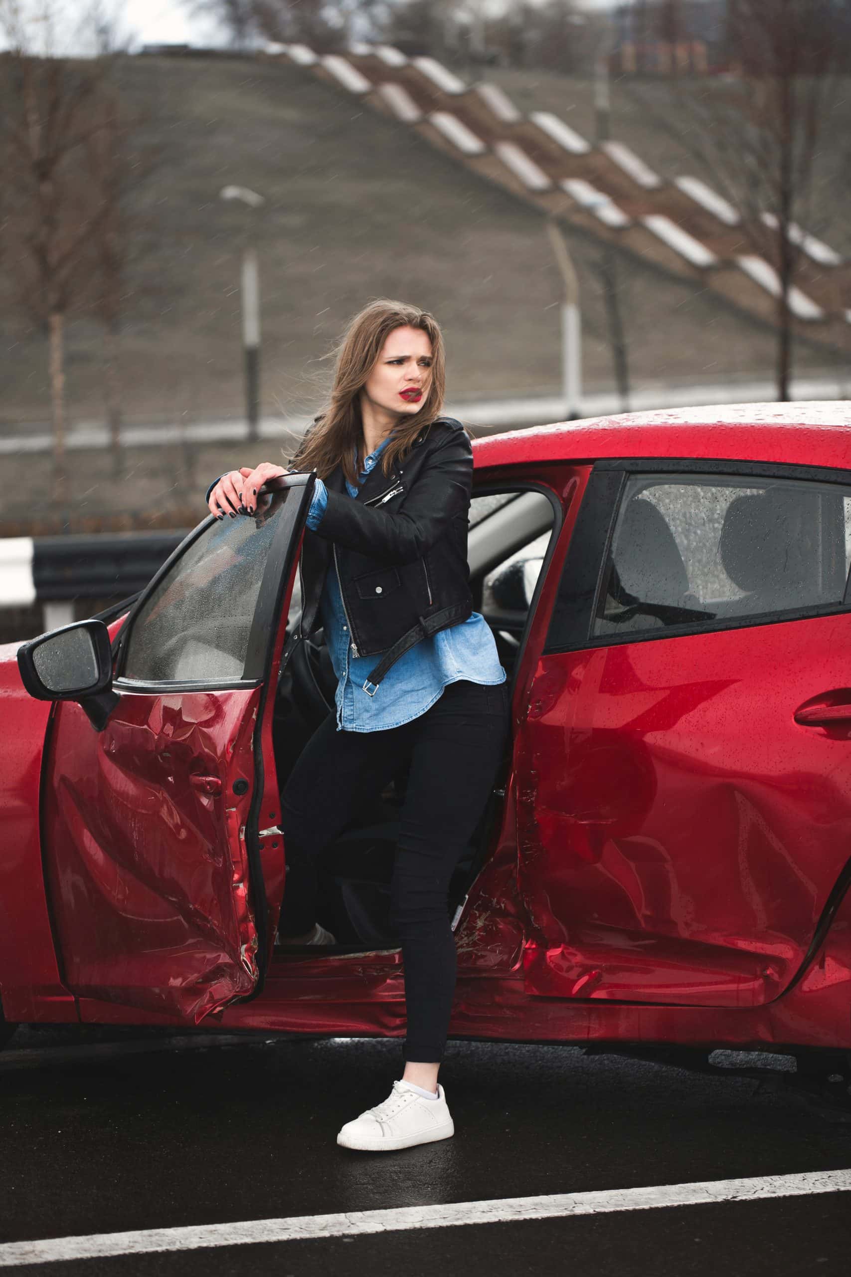 Woman exiting red damaged car on rainy day.