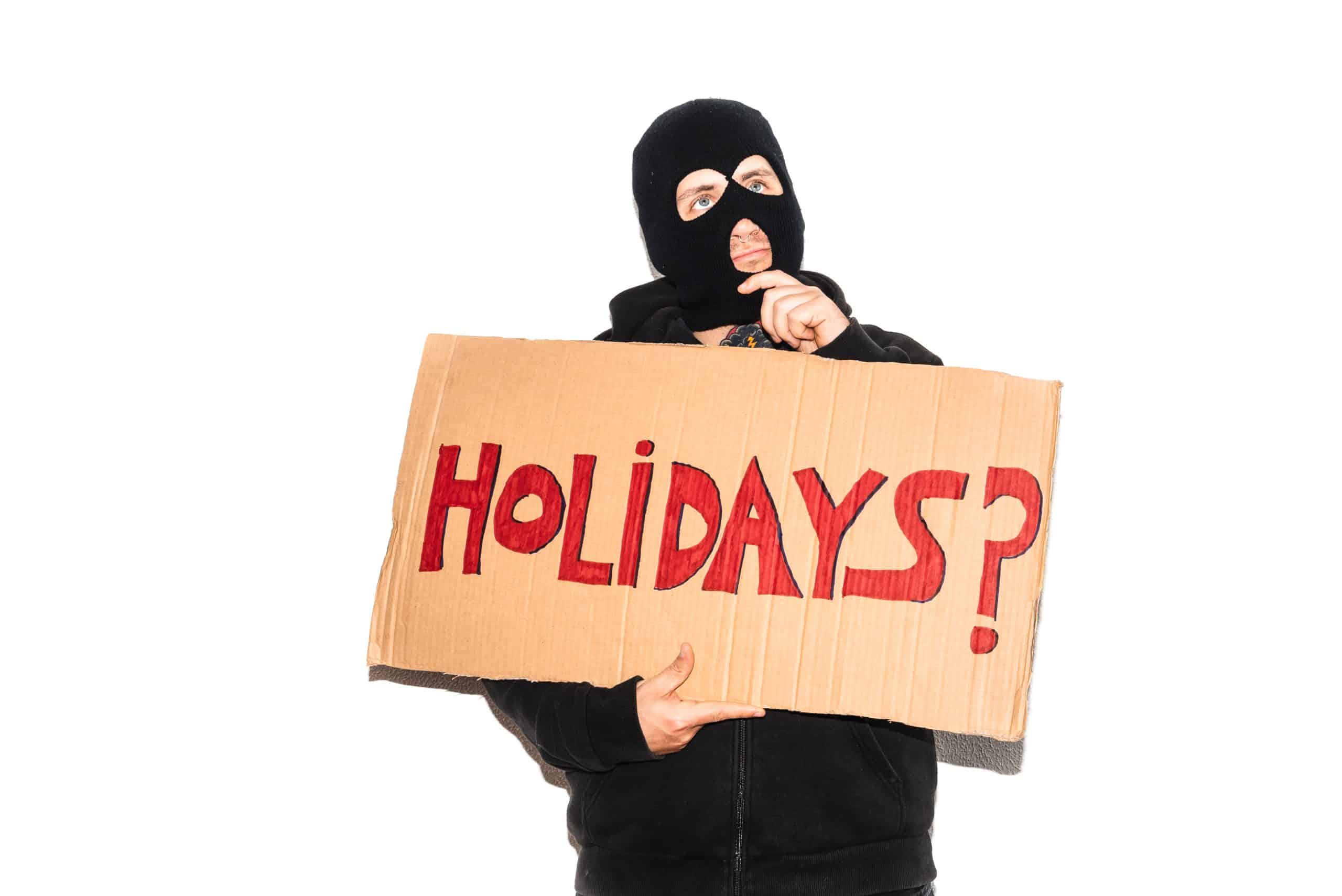 Person in ski mask holding 'Holidays?' sign.