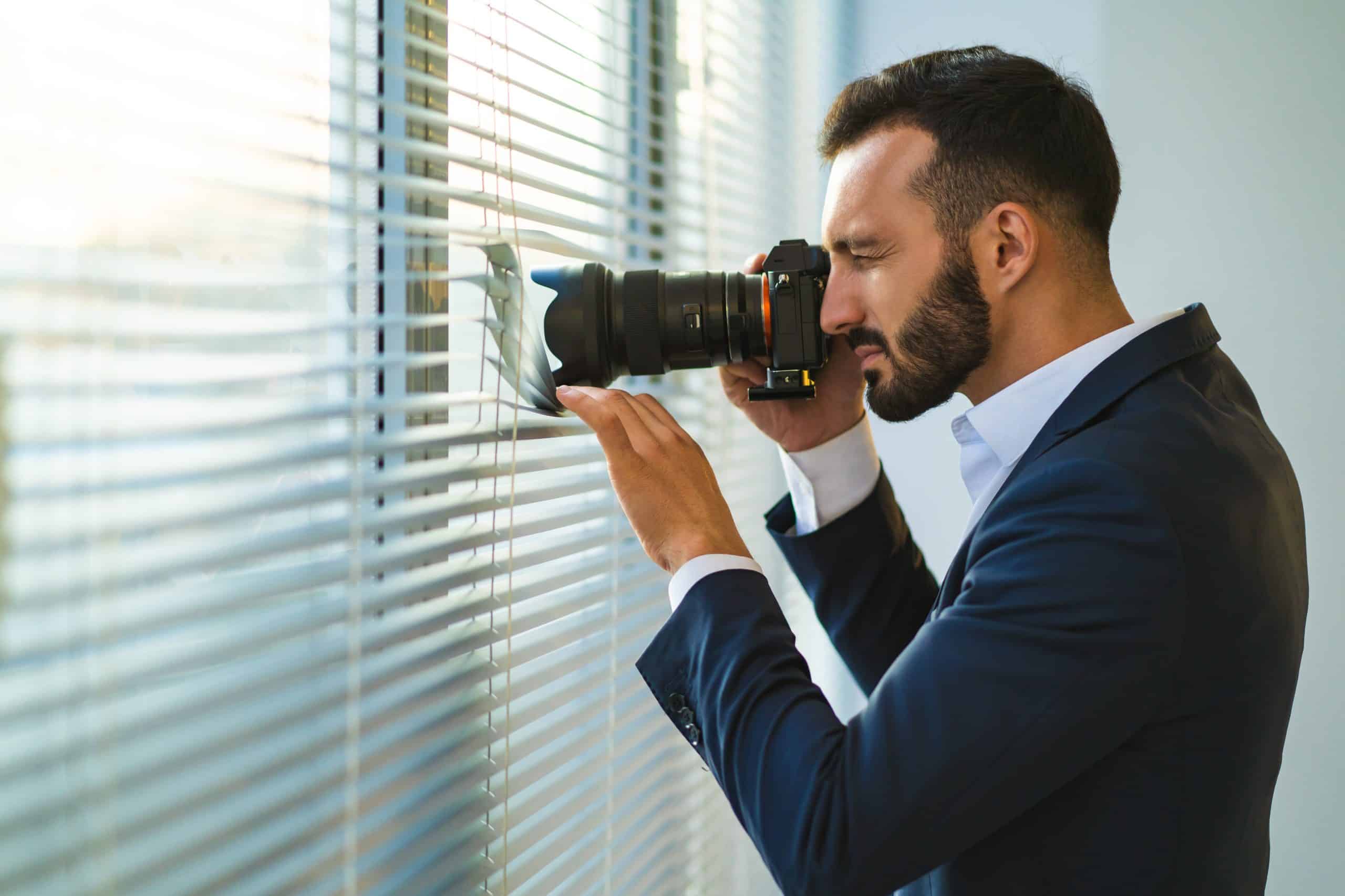 Man photographing through window blinds with camera.