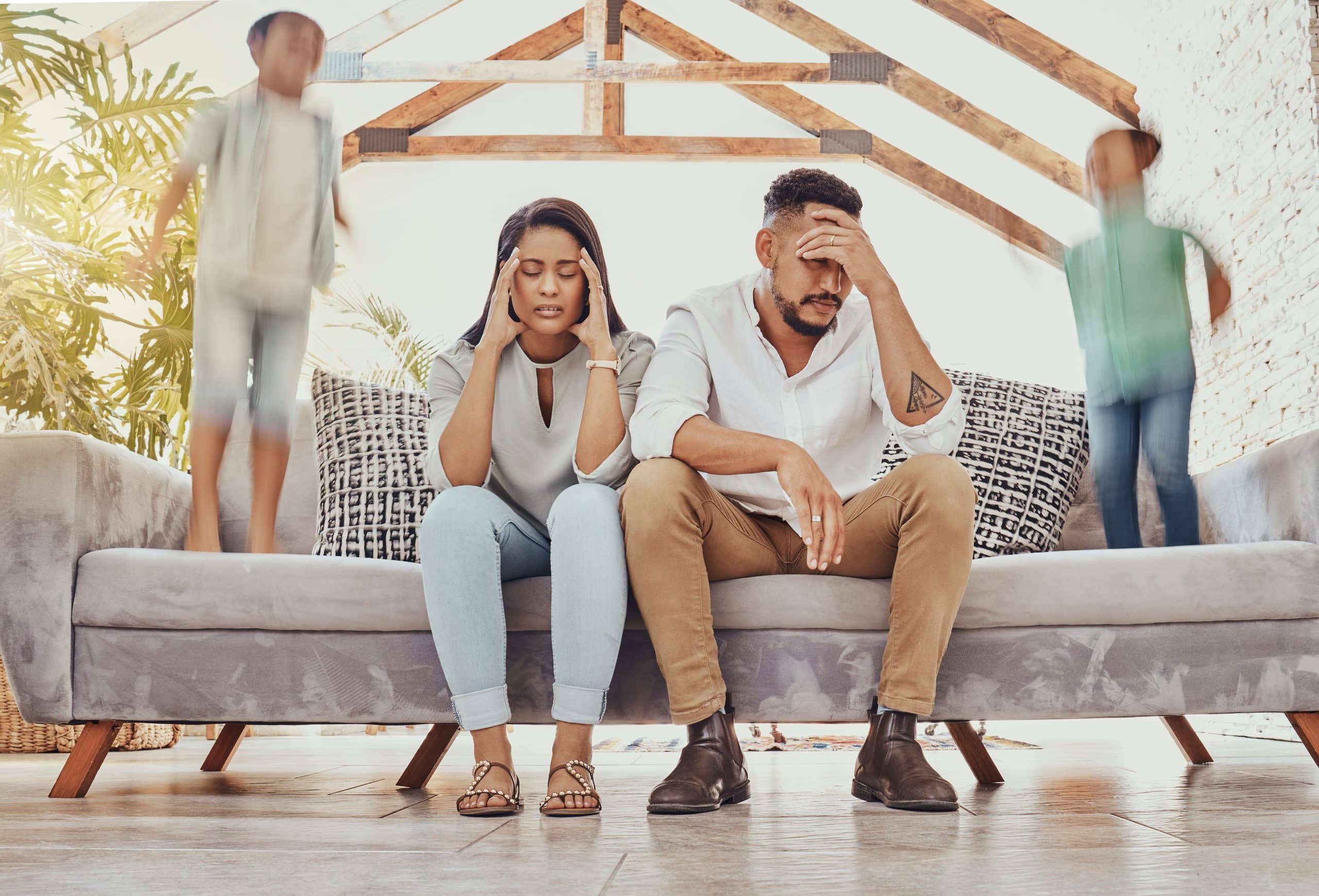 Stressed parents sitting while kids jump on couch.
